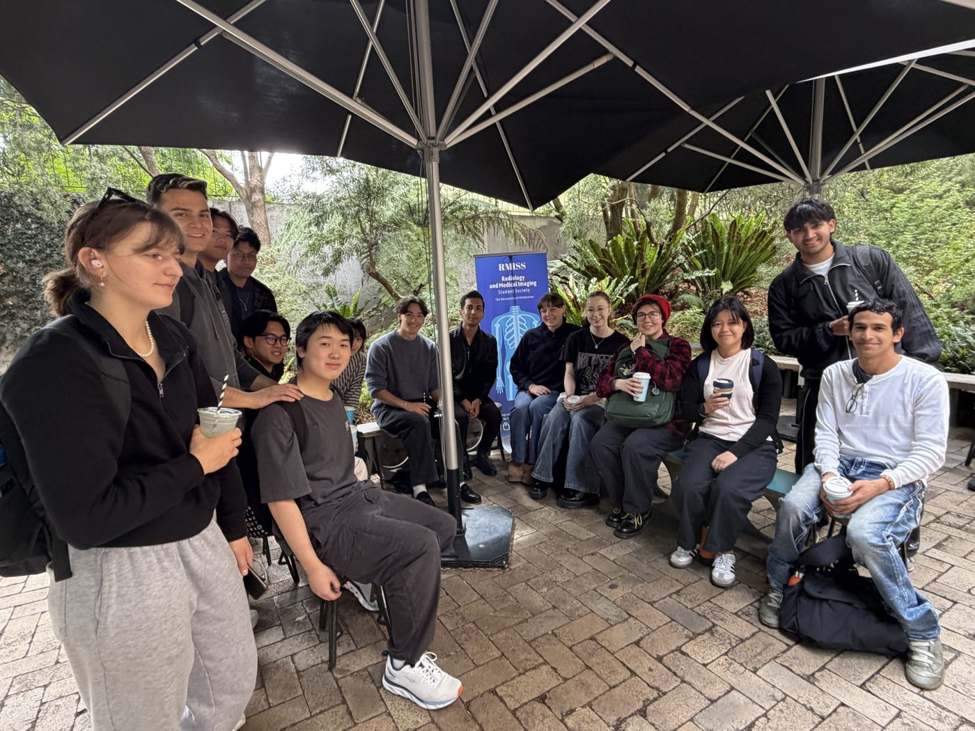 RMISS students gathered under large umbrellas beside the society banner